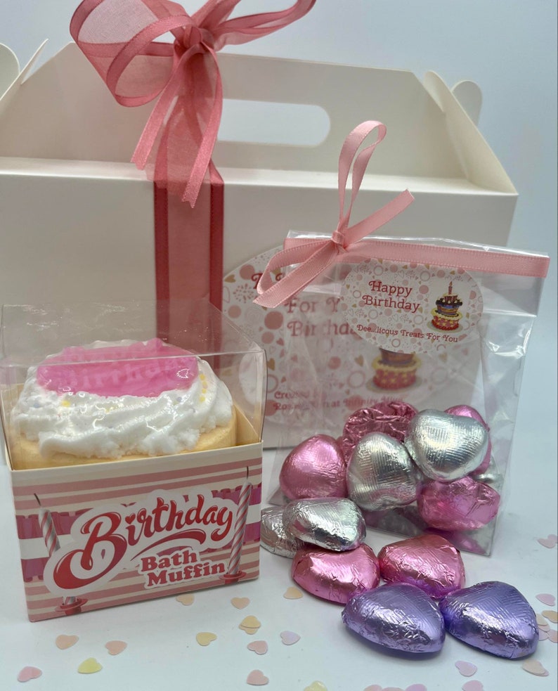 Puede incluir: Una caja de regalo blanca con una cinta rosa y una bolsa de pl&aacute;stico transparente llena de chocolates en forma de coraz&oacute;n rosa, plateado y morado. La caja tambi&eacute;n contiene una bomba de ba&ntilde;o rosa y blanca con forma de cupcake con las palabras "Birthday Bath Muffin" en la parte delantera.