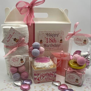 Puede incluir: Una caja de regalo rosa y blanca llena de productos de ba&ntilde;o para un 18 cumplea&ntilde;os. La caja incluye bombas de ba&ntilde;o, un muffin de ba&ntilde;o, sales de ba&ntilde;o y una toalla. La caja est&aacute; decorada con una cinta rosa y confeti.