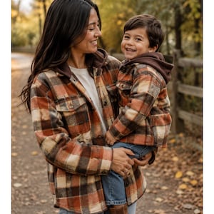 May include: Two matching long-sleeve button-up shirts featuring a plaid pattern in shades of orange, brown, cream, and black. Each shirt includes a dark brown hood, a front button closure, and two chest pockets with buttoned flaps on the front.