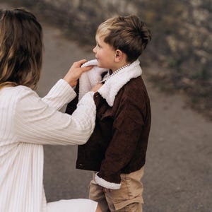 Puede incluir: Una chaqueta de pana marr&oacute;n oscuro con cuello de forro polar sherpa blanco y ribetes a juego en los pu&ntilde;os. La chaqueta tiene cierre frontal de botones y se lleva sobre una camisa a rayas, combinada con pantalones cargo de color tostado.