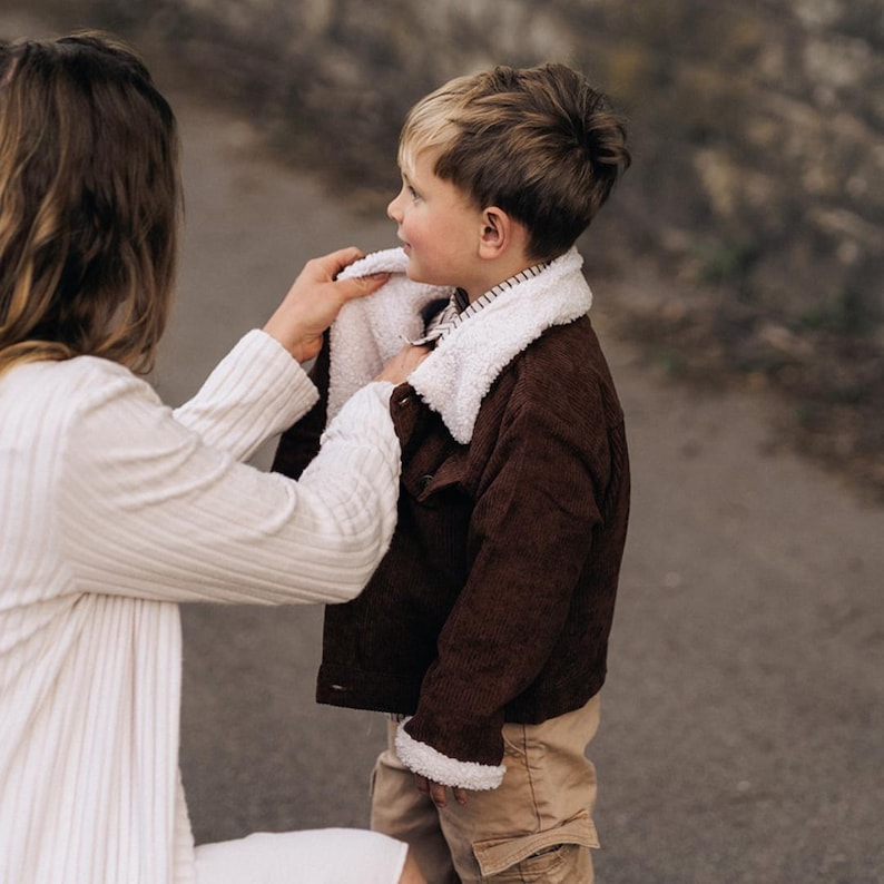 Puede incluir: Una chaqueta de pana marr&oacute;n oscuro con cuello de forro polar sherpa blanco y ribetes a juego en los pu&ntilde;os. La chaqueta tiene cierre frontal de botones y se lleva sobre una camisa a rayas, combinada con pantalones cargo de color tostado.