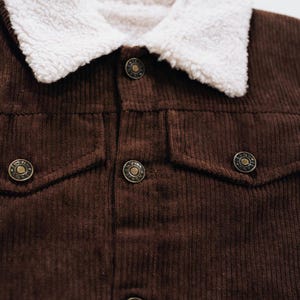Puede incluir: Una chaqueta de pana marr&oacute;n oscuro con cuello de sherpa blanco, cierre frontal de botones y dos bolsillos en el pecho con solapas abotonadas. Los botones de metal tienen texto grabado y un dise&ntilde;o circular con una apariencia texturizada.