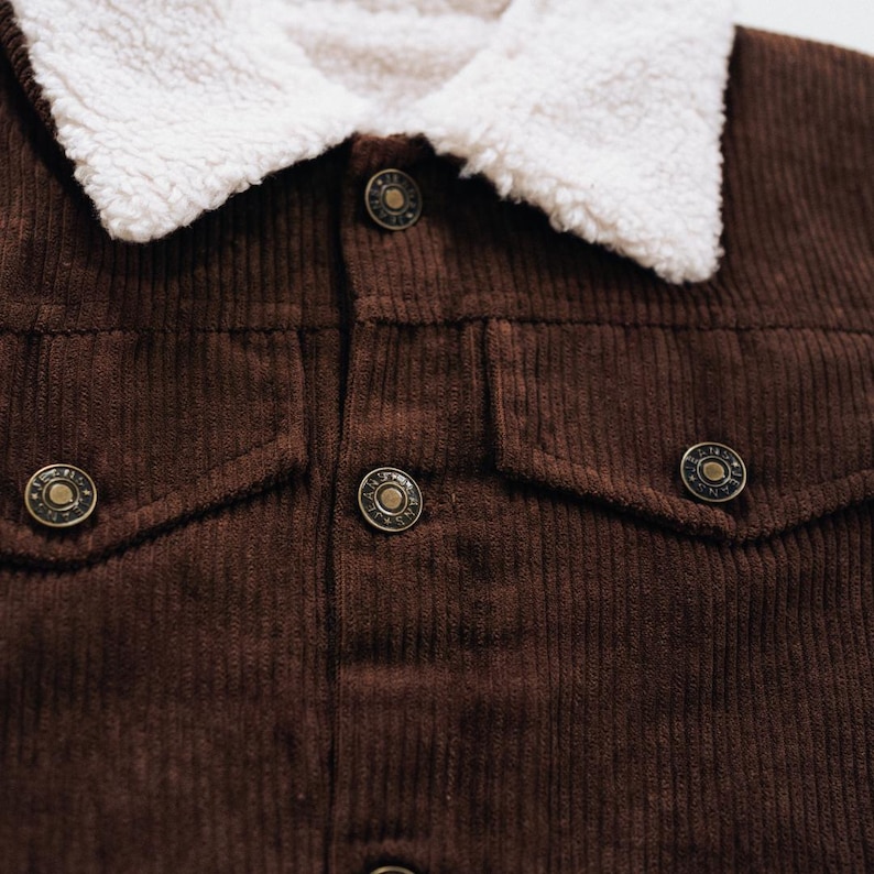 Puede incluir: Una chaqueta de pana marr&oacute;n oscuro con cuello de sherpa blanco, cierre frontal de botones y dos bolsillos en el pecho con solapas abotonadas. Los botones de metal tienen texto grabado y un dise&ntilde;o circular con una apariencia texturizada.