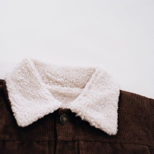 Puede incluir: Una chaqueta de pana marr&oacute;n oscuro con un cuello grueso de forro polar sherpa color crema. La prenda incluye un &uacute;nico bot&oacute;n met&aacute;lico visible en la parte delantera central, mostrando la textura de la pana y el forro polar suave y rizado.