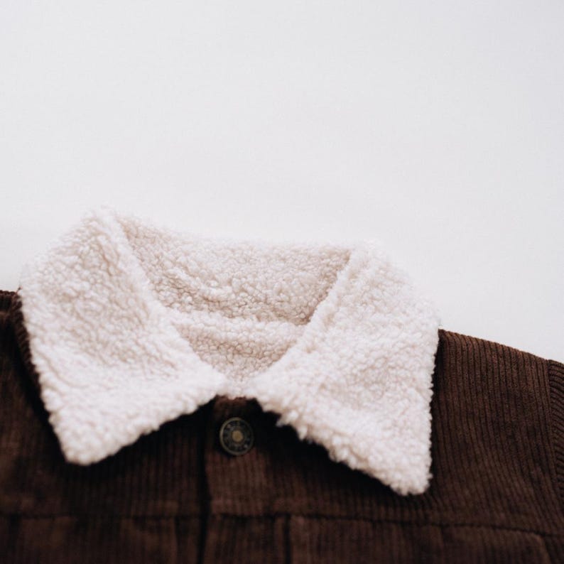 Puede incluir: Una chaqueta de pana marr&oacute;n oscuro con un cuello grueso de forro polar sherpa color crema. La prenda incluye un &uacute;nico bot&oacute;n met&aacute;lico visible en la parte delantera central, mostrando la textura de la pana y el forro polar suave y rizado.