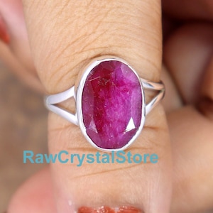 May include: A silver ring with a pink oval ruby gemstone. The ring is on a finger.
