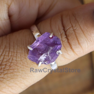 Op de afbeelding: Een zilveren ring met een grote, ruwe amethist edelsteen. De amethist is diep paars van kleur en heeft een ruwe, natuurlijke textuur. De ring wordt gedragen op een vinger met blauwe en roze stippen nagellak.