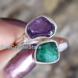 May include: A silver ring featuring two raw gemstones. The top stone is a purple amethyst and the bottom stone is a green emerald.