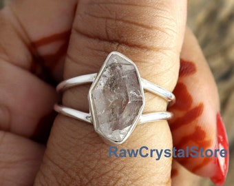 May include: A silver ring with a clear quartz crystal set in a double band design. The ring is worn on a person's thumb.