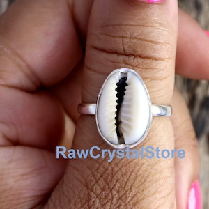 May include: A silver ring with a white cowrie shell set in the centre. The ring is on a person's thumb.