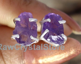 Puede incluir: Un par de pendientes de plata de ley con cristales de amatista en bruto. La amatista es de color morado intenso y los pendientes tienen un diseño sencillo.