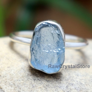 May include: A silver ring with a light blue aquamarine gemstone. The gemstone is raw and has a natural, irregular shape.