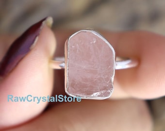May include: A silver ring with a raw rose quartz stone. The stone is a light pink color and has a natural, irregular shape.