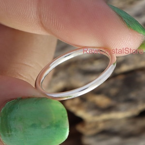 Puede incluir: Un anillo delgado de plata sostenido entre dedos con esmalte de uñas verde. El anillo es simple y brillante, reflejando la luz. El fondo es una superficie de madera natural. El texto "Raw Crystal Store" es visible.
