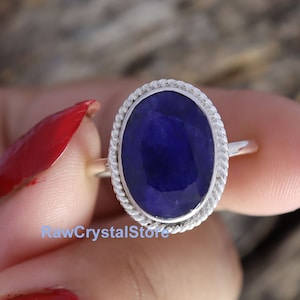 Peut inclure: Une bague en argent avec une pierre ovale bleu foncé sertie dans une bordure en forme de corde. La bague est sur un doigt avec du vernis à ongles rouge.