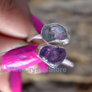 May include: Two raw crystal rings set in silver. The top ring features a gray-blue crystal, while the bottom ring features a purple amethyst crystal.