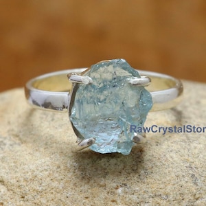 May include: A silver ring with a raw aquamarine gemstone. The gemstone is a light blue color and has a rough, natural shape. The ring is set in a simple silver band.