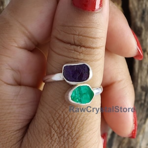May include: A silver ring with two raw gemstones. The top stone is a purple amethyst and the bottom stone is a green emerald. The ring is on a person's finger.