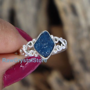 May include: A silver ring featuring a blue raw gemstone set in a diamond-shaped setting. The ring has a delicate, ornate design.