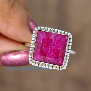 May include: A square-shaped ruby ring set in sterling silver. The ruby is a deep pink color and has a faceted cut. The ring has a beaded edge.