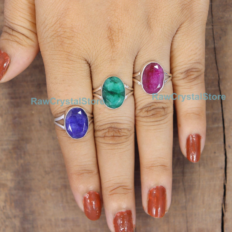 May include: Three sterling silver rings with oval shaped gemstones. The rings are on a person's fingers. The gemstones are a blue sapphire, a green emerald, and a red ruby.