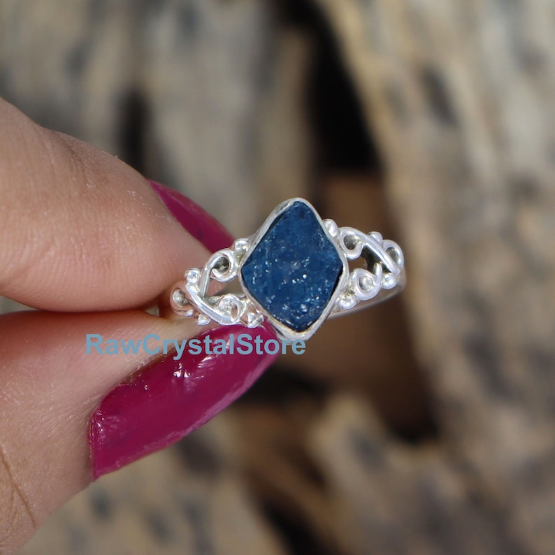 May include: A silver ring featuring a blue raw gemstone set in a diamond-shaped setting. The ring has a delicate, ornate design.