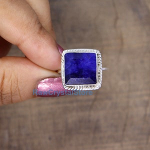 Pode incluir: Um anel de safira em forma quadrada engastado em prata de lei. A safira &eacute; de cor azul intensa e &eacute; rodeada por uma banda de prata com um design de corda.