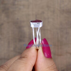 Peut inclure: Une bague en argent orn&eacute;e d'une pierre pr&eacute;cieuse rouge fonc&eacute;, de forme rectangulaire et brute. La bague a un fin anneau &agrave; double fil, et la pierre est sertie dans une lunette en argent. La bague est tenue par une personne avec du vernis &agrave; ongles rose.