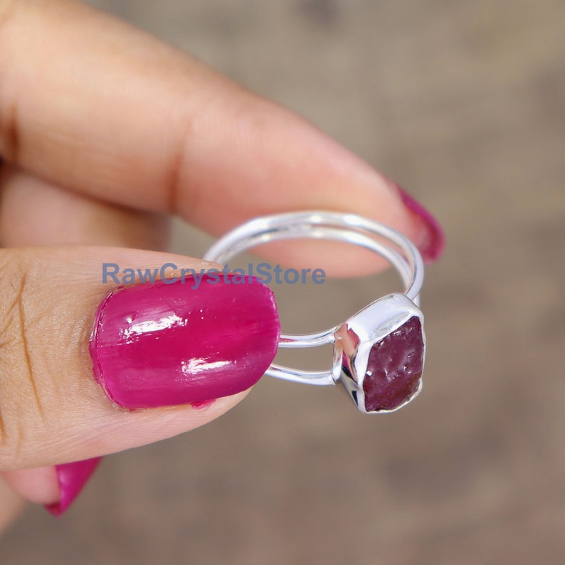 Peut inclure: Une bague en argent orn&eacute;e d'une pierre pr&eacute;cieuse brute en forme de rubis rectangulaire. La bague a un double anneau et le rubis est d'un rose-rouge profond. La bague est tenue par une personne avec du vernis &agrave; ongles rose.