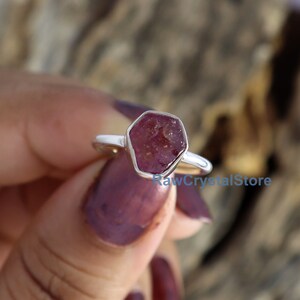 May include: A silver ring featuring a raw, faceted ruby gemstone. The ruby is a deep reddish-pink color and is set in a simple, polished silver band. The ring has a natural, organic aesthetic.