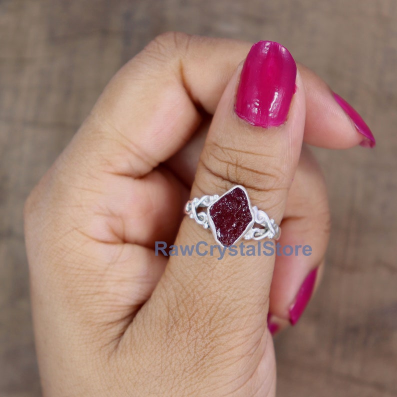 May include: A silver ring featuring a dark red, raw ruby gemstone set in a diamond-shaped bezel. The ring has a delicate, floral design on the band.