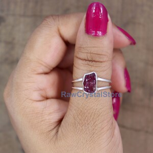 Peut inclure: Une bague en argent orn&eacute;e d'une pierre pr&eacute;cieuse brute, rectangulaire, rouge fonc&eacute;. La bague a un design &agrave; double bande et la pierre est sertie dans une lunette. Le texte "RawCrystalStore" est visible.