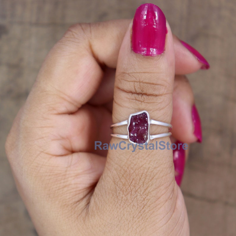 Peut inclure: Une bague en argent orn&eacute;e d'une pierre pr&eacute;cieuse brute, rectangulaire, rouge fonc&eacute;. La bague a un design &agrave; double bande et la pierre est sertie dans une lunette. Le texte "RawCrystalStore" est visible.