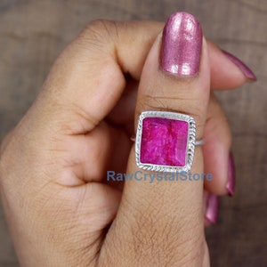 May include: A silver ring with a square pink ruby gemstone. The ring is set in a silver bezel with a braided edge. The ring is on a person's finger.