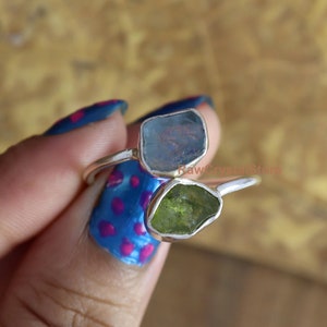May include: A silver ring with two raw gemstones. The larger stone is a light blue aquamarine, and the smaller stone is a green peridot. The ring is on a finger with blue and pink nail polish.