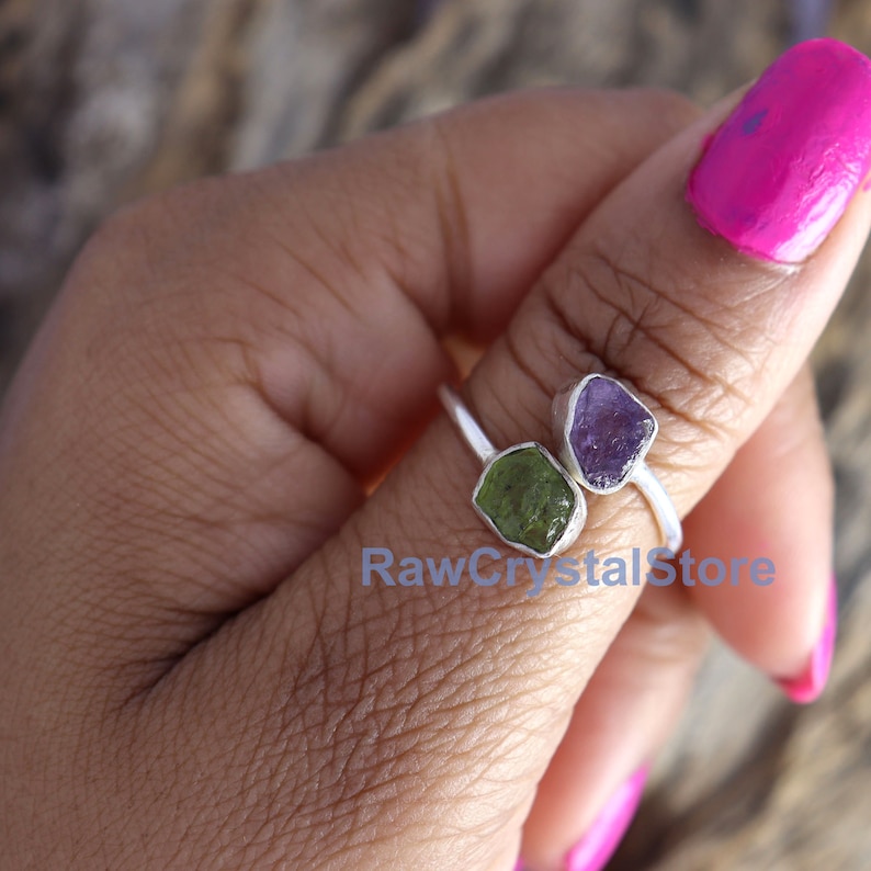 May include: A silver ring with two raw gemstones. The ring features a green peridot and a purple amethyst. The ring is on a person's finger.