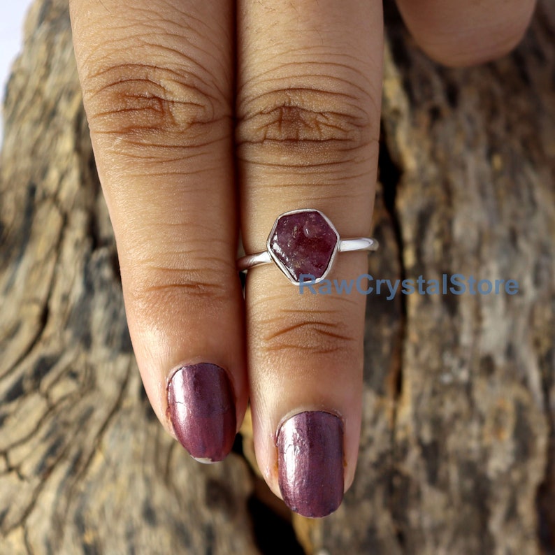 May include: A silver ring featuring a hexagonal, raw ruby gemstone. The ruby is a deep red color and set in a simple, polished silver band. The ring is shown on a finger with dark red nail polish, against a wooden background.
