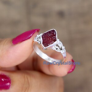 May include: A silver ring featuring a red, raw crystal set in a delicate, ornate design. The ring is on a finger with pink nail polish.