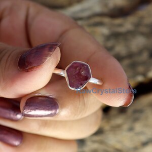 May include: A silver ring featuring a raw, faceted, dark red gemstone. The gemstone is set in a simple, polished silver band. The ring is held by a person with maroon nail polish. The text "RawCrystalStore" is visible.