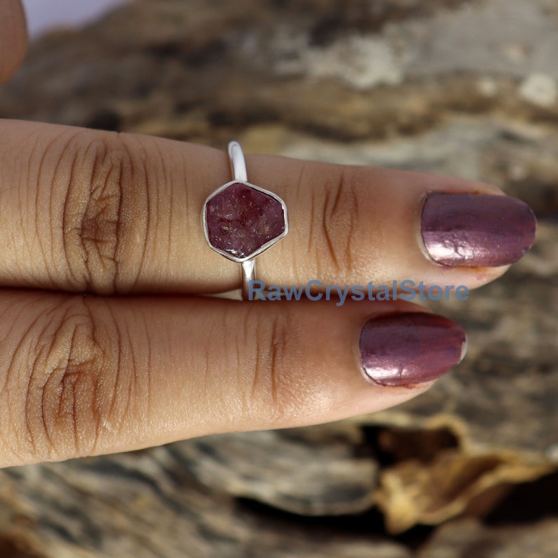 May include: A silver ring featuring a raw, hexagonal-shaped, deep red gemstone. The ring's band is thin and polished, and the gemstone is set in a silver bezel. The ring is displayed on a finger. The text "RawCrystalStore" is visible.