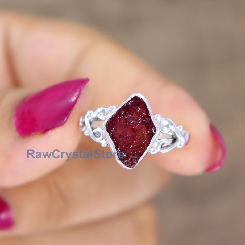 May include: A silver ring featuring a raw ruby gemstone set in a diamond-shaped bezel. The ring has a delicate, ornate design.