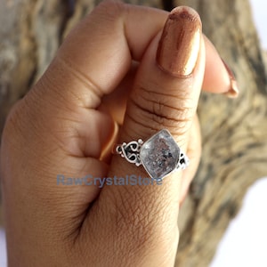 May include: A silver ring featuring a clear, faceted, square-shaped gemstone. The ring has a delicate, ornate design.