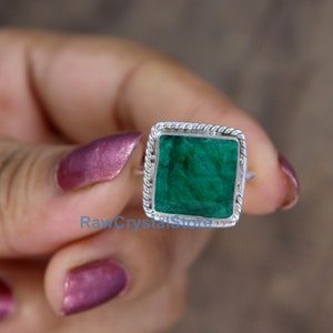Pode incluir: Um anel de prata com uma pedra preciosa quadrada de cor verde esmeralda engastada em uma moldura de prata. O anel est&aacute; em um dedo.