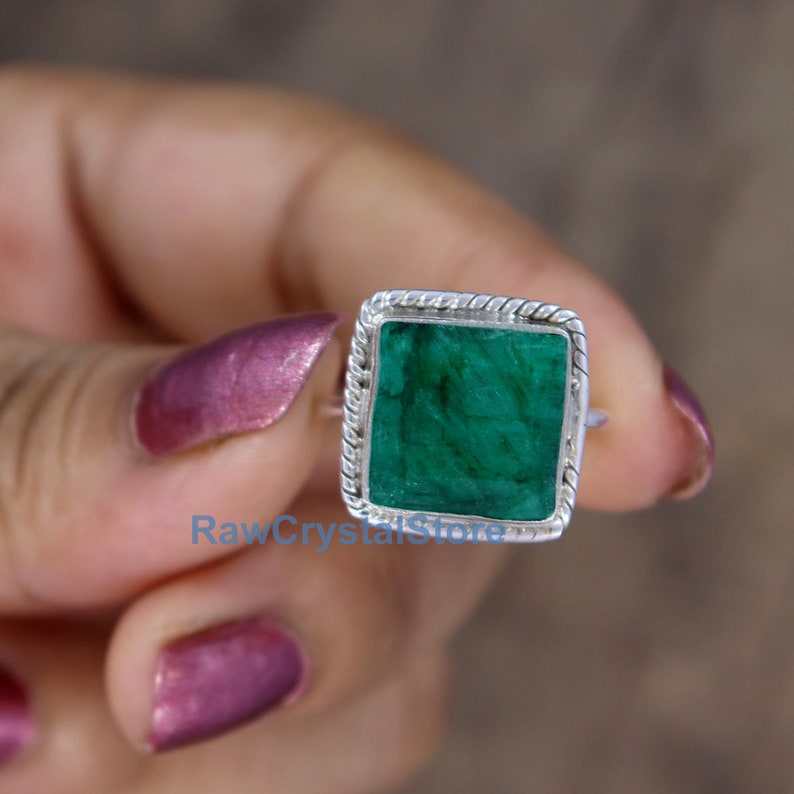 Pode incluir: Um anel de prata com uma pedra preciosa quadrada de cor verde esmeralda engastada em uma moldura de prata. O anel est&aacute; em um dedo.