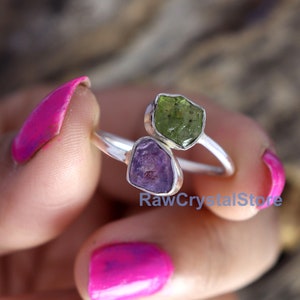 May include: A silver ring with two raw gemstones. The larger stone is a green peridot and the smaller stone is a purple amethyst.