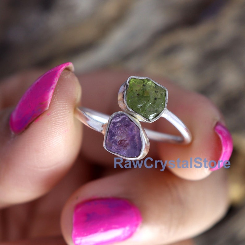 May include: A silver ring with two raw gemstones. The larger stone is a green peridot and the smaller stone is a purple amethyst.