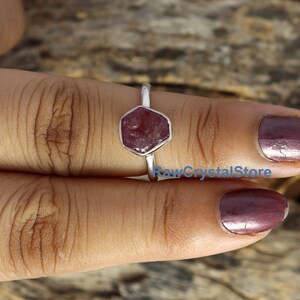 May include: A silver ring featuring a hexagonal, raw ruby gemstone. The ruby is a deep reddish-pink color. The ring is displayed on a person's finger, with the text "RawCrystalStore" visible in the image.
