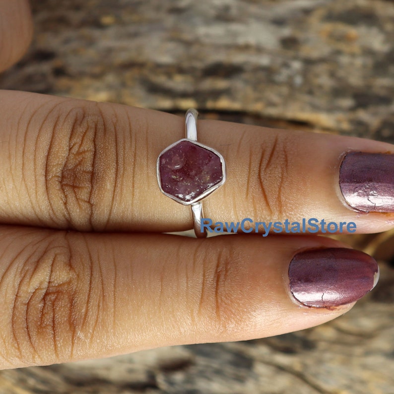 May include: A silver ring featuring a hexagonal, raw ruby gemstone. The ruby is a deep reddish-pink color. The ring is displayed on a person's finger, with the text "RawCrystalStore" visible in the image.