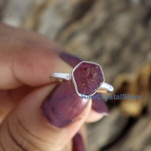 May include: A silver ring featuring a raw, faceted ruby gemstone. The ruby is a deep reddish-pink color and is set in a simple, polished silver band. The ring is shown against a blurred wooden background.