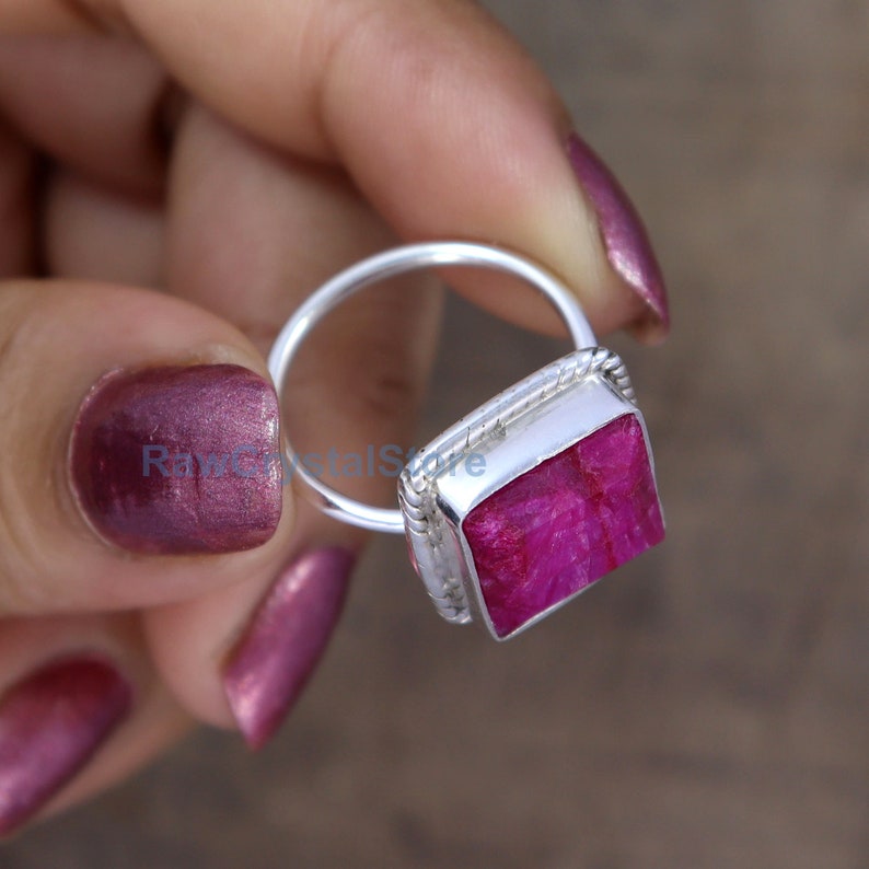 Pode incluir: Um anel de prata com uma pedra preciosa de rubi rosa de forma quadrada. O anel tem uma banda tran&ccedil;ada.